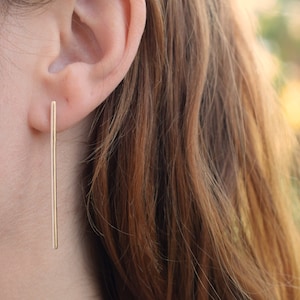 May include: A close-up of a person's ear with a long, thin gold bar earring. The earring is simple and elegant.