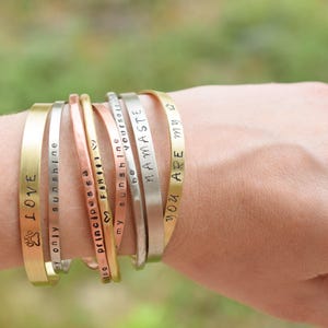 May include: A stack of gold, silver, and copper bracelets with various engraved messages, including "LOVE", "my only sunshine", "principessa", "FAMILY", "SunShine", "be yourself", "NAMASTE", and "YOU ARE MY".