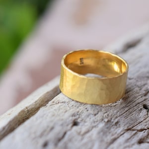 Può includere: Un anello a fascia larga in oro con una texture martellata. L'anello è appoggiato su un pezzo di legno di deriva.