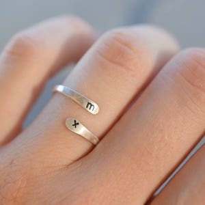Op de afbeelding: Een zilverkleurige wikkelring met de letters "m" en "x" op de uiteinden. De ring wordt om een vinger gedragen, waarbij de uiteinden elkaar overlappen. De ring heeft een eenvoudig, minimalistisch ontwerp.