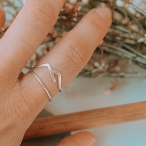 May include: A silver ring featuring a mountain range design. The ring is worn on a finger and is stacked with a thin silver band.