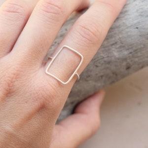 May include: A silver ring with a rectangular shape. The ring is worn on a finger.