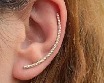 Puede incluir: Un ear cuff dorado con un acabado texturizado y martillado. El diseño curvado sigue la forma natural de la oreja, creando un look moderno y minimalista. Una adición única a cualquier colección de joyas.