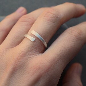Op de afbeelding: Een zilveren ring met een uniek ontwerp met twee dunne banden die elkaar kruisen. De ring wordt aan een vinger gedragen.