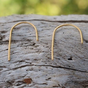 Op de afbeelding: Een paar gouden, minimalistische oorbellen in de vorm van een boog. De oorbellen liggen op een houten oppervlak.