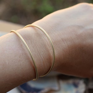 Può includere: Due braccialetti rigidi dorati impilati sul polso di una persona. I braccialetti sono semplici e minimalisti nel design.