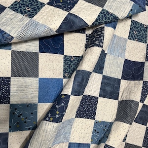 May include: A patchwork quilt in shades of blue and white. The quilt features a variety of patterned squares, including polka dots, geometric shapes, and abstract designs. The quilt is folded, showing the different patterns and textures.