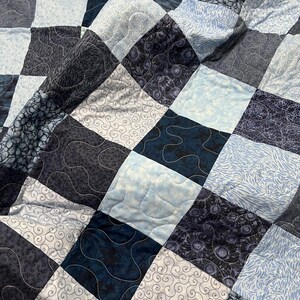 May include: A handmade quilt with a patchwork design. The quilt features squares in various shades of blue, gray, and white, with different patterns including swirls, floral prints, and solid colors. The quilt is machine quilted with white thread.