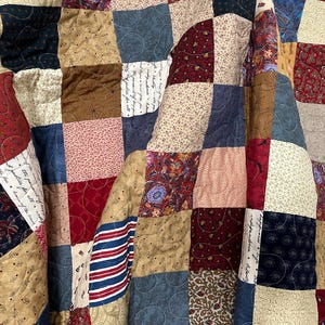 May include: A patchwork quilt with a variety of fabrics in shades of red, blue, tan, and cream. The quilt features a mix of floral, striped, and patterned fabrics, creating a cozy and rustic aesthetic. The quilt is likely handmade.