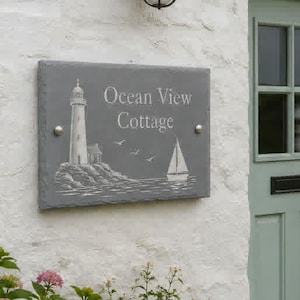 May include: A rectangular grey slate sign mounted on a white stone wall, featuring an engraved lighthouse, a sailboat, and the text Ocean View Cottage. The sign is positioned next to a sage green door with a black handle and a black outdoor lamp.