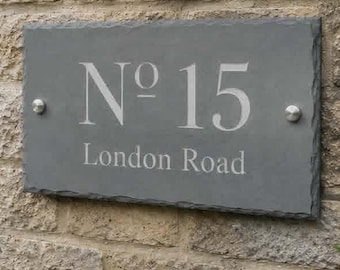 May include: A rectangular grey slate house sign with rough, chiseled edges, mounted on a tan stone wall. The sign features the engraved white text "No 15" above "London Road" and is secured by two silver-colored metal bolts on the left and right sides.