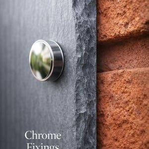 May include: A dark gray slate plaque with a rough, chiseled edge and a circular, reflective chrome fixing mounted on the surface. The text "Chrome Fixings" is printed in white at the bottom left of the slate plaque, which is positioned against a brick wall.