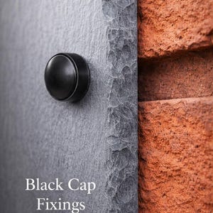 May include: A dark gray slate plaque with a rough, chiseled edge mounted against a textured red brick wall. A circular black cap fixing is attached to the front surface. The text Black Cap Fixings is printed in white on the lower left side.