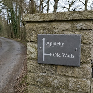 May include: A rectangular dark gray slate sign mounted on a stone pillar with white engraved text and arrows. The sign features an upward-pointing arrow next to the word Appleby and a right-pointing arrow next to the words Old Walls.