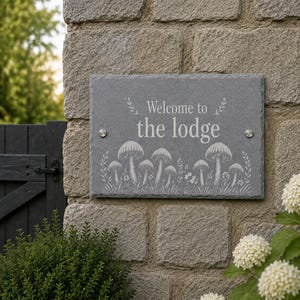 May include: A rectangular grey slate sign with rough, natural edges. The text "Welcome to the lodge" is engraved in white, centred above a detailed white illustration of various mushrooms and small plants. Two silver metal bolts secure the sign.
