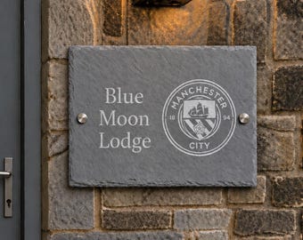 May include: A rectangular gray slate sign mounted on a stone wall with two silver metal fasteners. The sign features the engraved text Blue Moon Lodge and the Manchester City football club logo, including a ship and the numbers 18 and 94.