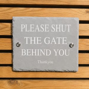 May include: A rectangular grey slate sign with rough, natural edges. The text in white reads: PLEASE SHUT THE GATE BEHIND YOU, followed by Thankyou. Two silver-colored metal circular fasteners are positioned on the left and right sides of the sign.
