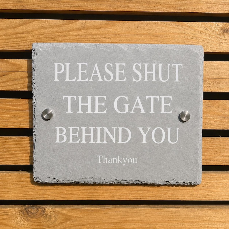 May include: A rectangular grey slate sign with rough, natural edges. The text in white reads: PLEASE SHUT THE GATE BEHIND YOU, followed by Thankyou. Two silver-colored metal circular fasteners are positioned on the left and right sides of the sign.