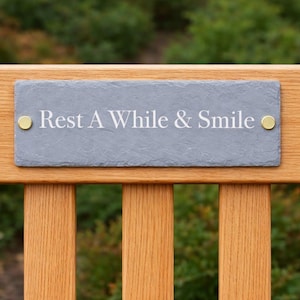 May include: A wooden bench with a grey slate plaque inscribed with the words "Rest A While & Smile" in white. The plaque is fixed with two gold-coloured screws. The bench has a natural wood finish.