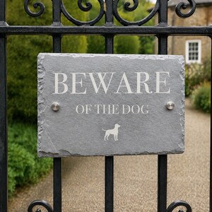 May include: A rectangular gray slate sign with rough, natural edges mounted on a black metal gate. The sign features the white engraved text BEWARE OF THE DOG and a small white silhouette of a dog. Two silver circular metal bolts secure the sign.