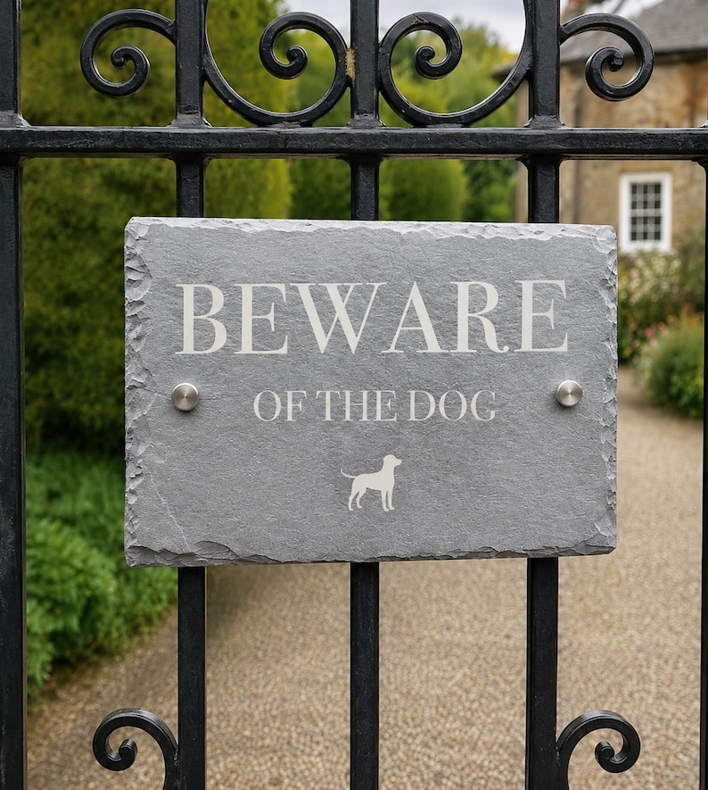 May include: A rectangular gray slate sign with rough, natural edges mounted on a black metal gate. The sign features the white engraved text BEWARE OF THE DOG and a small white silhouette of a dog. Two silver circular metal bolts secure the sign.