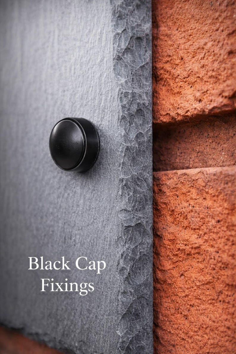 May include: A dark gray slate plaque with a rough, chiseled edge featuring a smooth, circular black cap fixing. The text Black Cap Fixings is printed in white on the lower left side of the slate surface, which is mounted against a red brick wall.