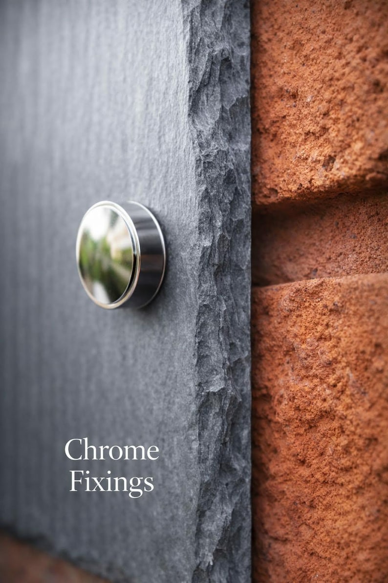 May include: A dark gray slate plaque with a rough, textured edge and a polished, circular chrome fixing attached to the surface. The text "Chrome Fixings" is printed in white on the lower left side of the slate, which is mounted against a red brick wall.