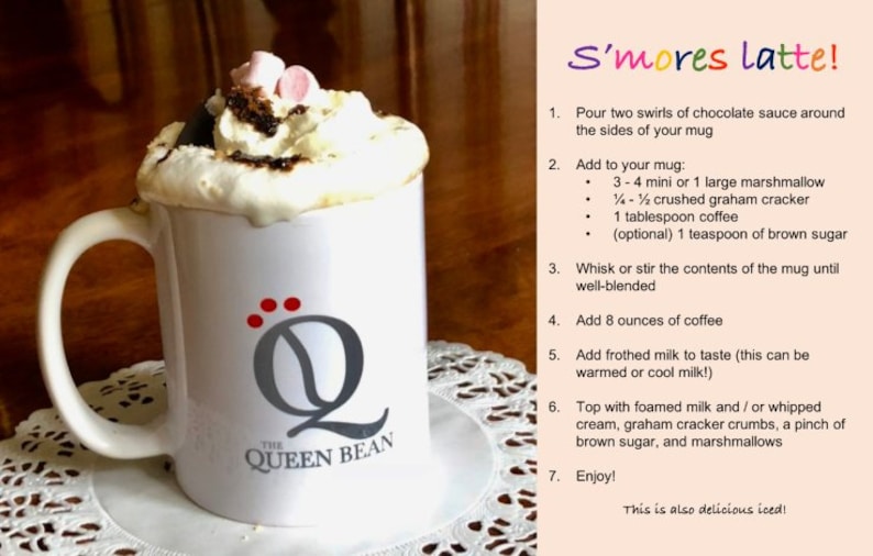 May include: A white mug with a gray "Q" logo and the text "The Queen Bean" is filled with a s'mores latte. The latte is topped with whipped cream, chocolate sauce, marshmallows, and graham cracker crumbs. The recipe for the s'mores latte is written on a pink background.