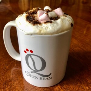 S'mores coffee in a Queen Bean mug topped with whipped cream, marshmallows, and brown sugar