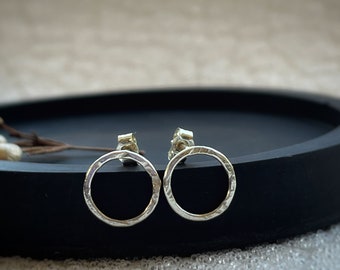 May include: A pair of silver circle stud earrings with a hammered texture. The earrings are on a black surface.