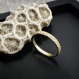 May include: A gold-colored ring with a textured surface sits on a black surface next to a piece of white coral. The ring's band has a detailed, hammered appearance. The coral has a porous, honeycomb-like structure.