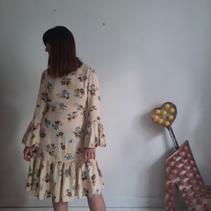 May include: A woman wearing a long-sleeved, cream-colored dress with a floral print. The dress has a ruffled hem and bell sleeves. The woman is standing in front of a white wall with a heart-shaped sign behind her.