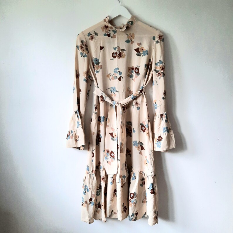 May include: A beige dress with a floral print and a tie waist. The dress has long sleeves with bell cuffs and a ruffled hem.
