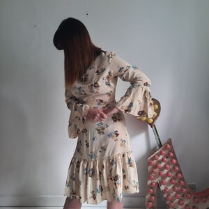 May include: A woman wearing a beige dress with a floral print and long sleeves. The dress has a ruffled hem and a cinched waist. The sleeves are bell-shaped and have a floral print.
