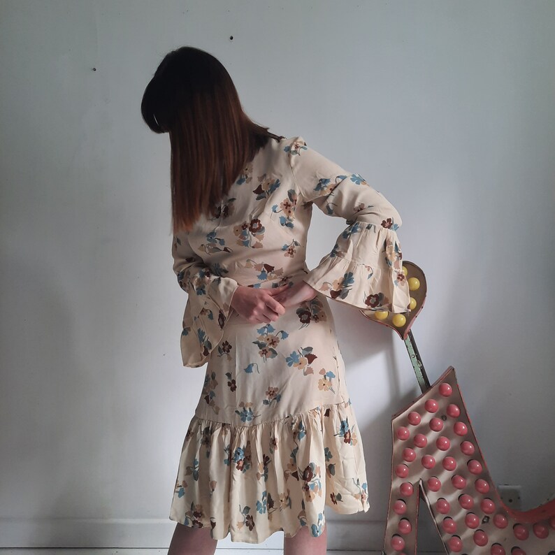 May include: A woman wearing a beige dress with a floral print and long sleeves. The dress has a ruffled hem and a cinched waist. The sleeves are bell-shaped and have a floral print.
