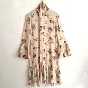 May include: A cream-colored dress with a floral print in shades of brown, blue, and white. The dress has a tie at the neck and bell sleeves.