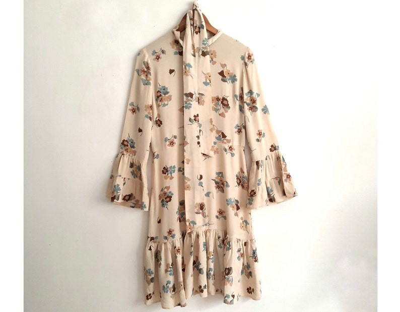 May include: A cream-colored dress with a floral print in shades of brown, blue, and white. The dress has a tie at the neck and bell sleeves.