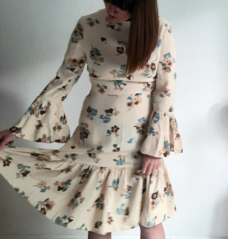 May include: A cream-colored midi dress with a floral print featuring blue, brown, and beige flowers. The dress has long sleeves with bell cuffs and a ruffled hem.