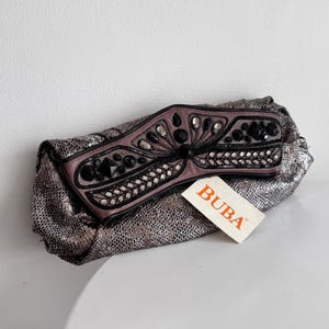 Könnte beinhalten: Eine silberne Clutch mit Schlangenmuster. Die obere Klappe ist mauvefarben mit schwarzen und silbernen Verzierungen. Die Clutch hat ein Etikett mit der Aufschrift "BUBA".