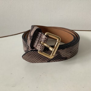 May include: A coiled belt with a snakeskin pattern in shades of brown and tan. The belt features a gold-coloured square buckle and a black edge. The interior of the belt is a light brown colour.