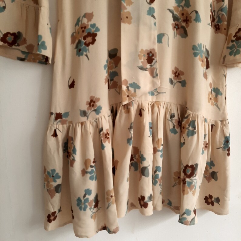 May include: A cream-colored dress with a floral print in shades of brown, blue, and green. The dress has a ruffled hem and a tie at the neck.