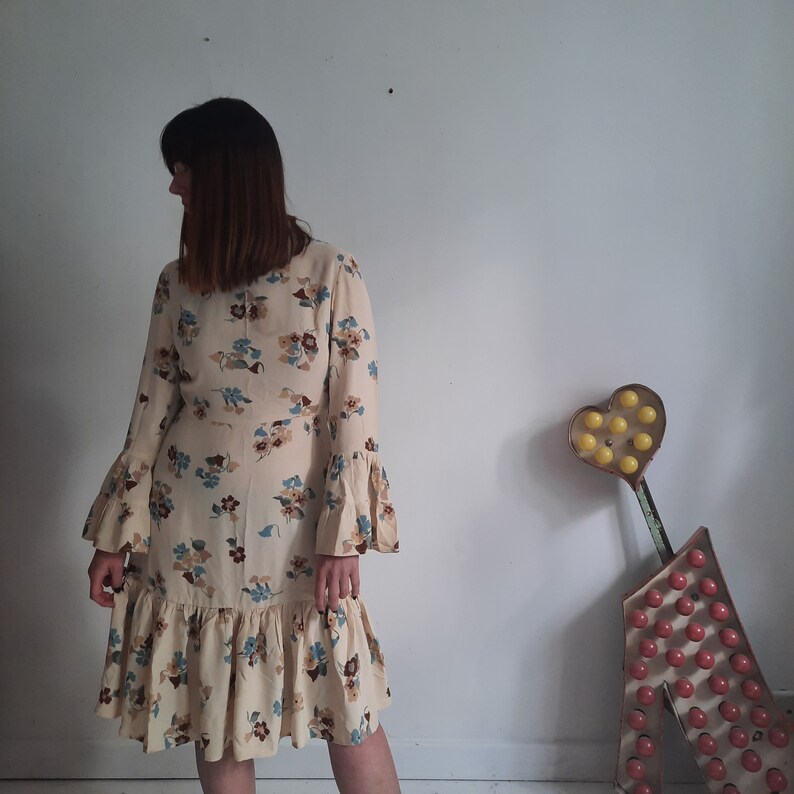 May include: A woman wearing a long-sleeved, cream-colored dress with a floral print. The dress has a ruffled hem and bell sleeves. The woman is standing in front of a white wall with a heart-shaped sign behind her.