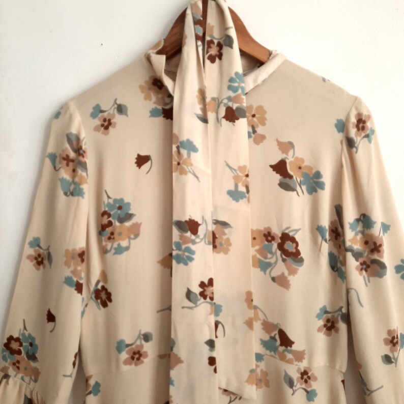 May include: A cream-colored long-sleeved dress with a floral print in shades of brown, blue, and green. The dress has a high neck and a tie at the neck.