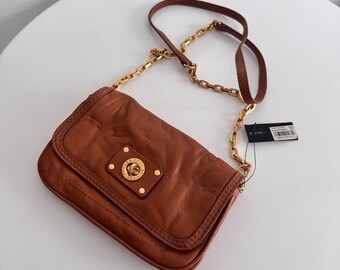 May include: A brown leather crossbody bag with a gold chain strap. The bag has a rectangular shape with a flap closure and a gold logo on the front. The bag is approximately 23 cm wide and 15 cm tall.