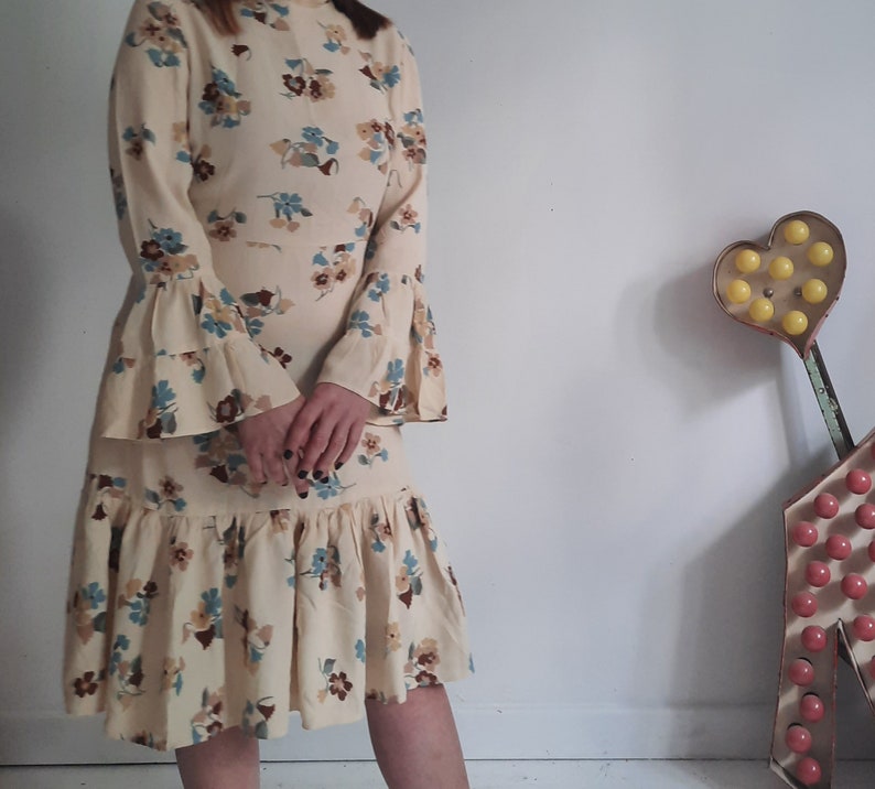 May include: A cream-colored dress with a floral print and bell sleeves. The dress has a tiered skirt and a fitted bodice.