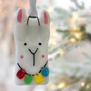 May include: White ceramic llama ornament with pink ears and a black face. The llama is wearing a string of colorful lights around its neck.
