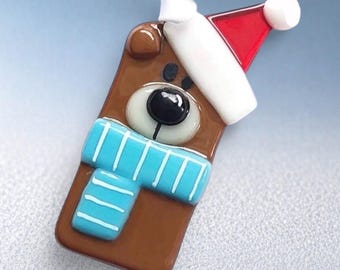 May include: A brown bear-shaped Christmas ornament with a Santa hat and a blue scarf. The ornament has a white ribbon for hanging. The bear has a black nose and a white and light brown face.