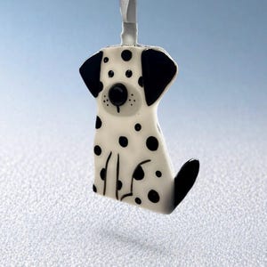 May include: A white glass ornament shaped like a sitting Dalmatian dog with black spots, black ears, and a black tail. The ornament features a round black nose and a white ribbon loop at the top for hanging, displaying a smooth, glossy finish.
