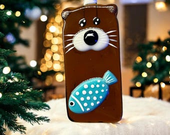 May include: A brown glass ornament shaped like an otter holding a blue and white fish with white polka dots. The ornament has a white string for hanging.