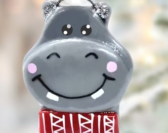 May include: A gray ceramic Christmas ornament shaped like a smiling hippopotamus wearing a red and white striped scarf.